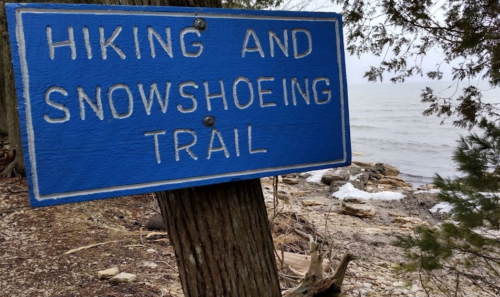 Blue sign reading "Hiking and Snowshoeing Trail" near a rocky shoreline with water in the background.