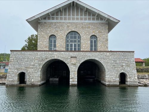 A stone building with large arched openings sits by a calm body of water, surrounded by greenery and other structures.