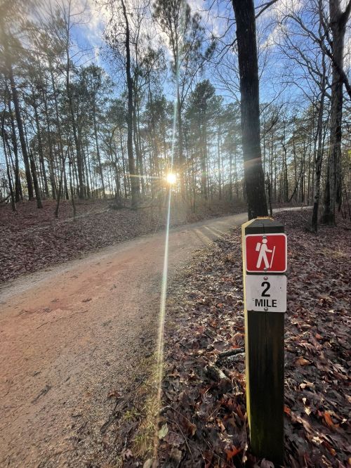Trail sign indicating 2 miles to go, with a sun setting through trees in a wooded area.
