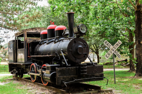 A vintage steam locomotive displayed near a railroad crossing sign, surrounded by trees.