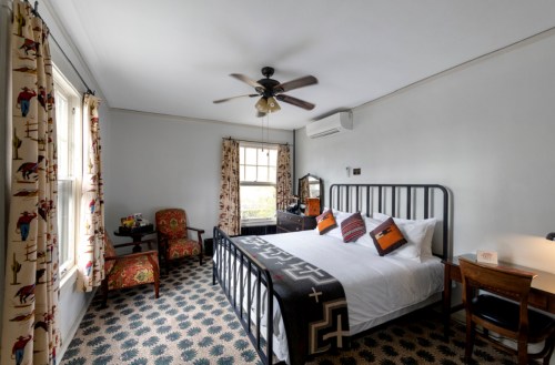 Cozy hotel room featuring a bed, patterned carpet, armchair, and decorative curtains with a fan overhead.