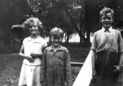 Three children stand outdoors, smiling, with trees and a building in the background. The photo is in black and white.