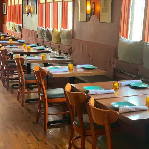 A cozy restaurant interior with wooden tables, chairs, and soft seating along the walls, illuminated by warm lighting.