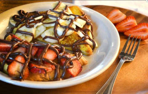 A plate of French toast topped with banana slices, strawberries, and drizzled chocolate sauce, with a fork on the side.