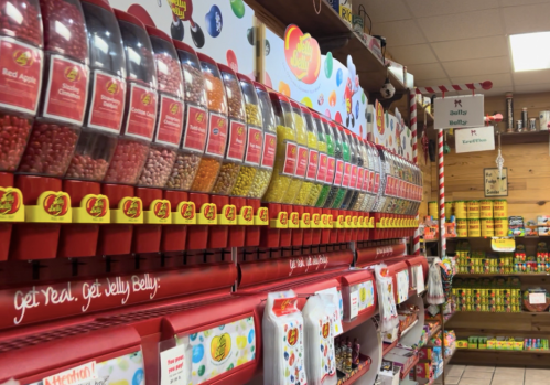A colorful display of jelly bean dispensers in a candy shop, with various candies and snacks on shelves in the background.