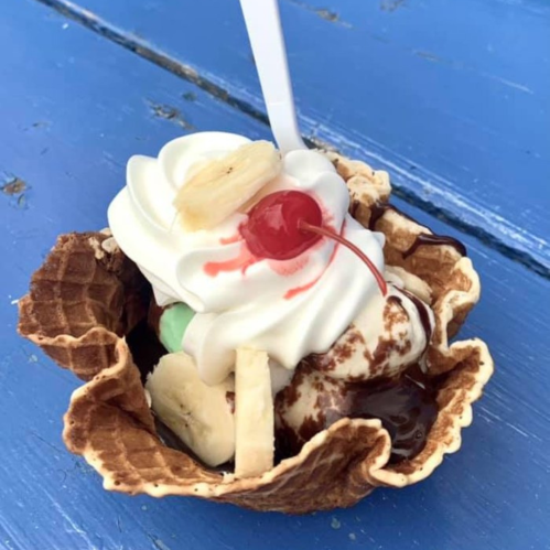A waffle bowl filled with ice cream, topped with whipped cream, a cherry, and banana slices on a blue table.