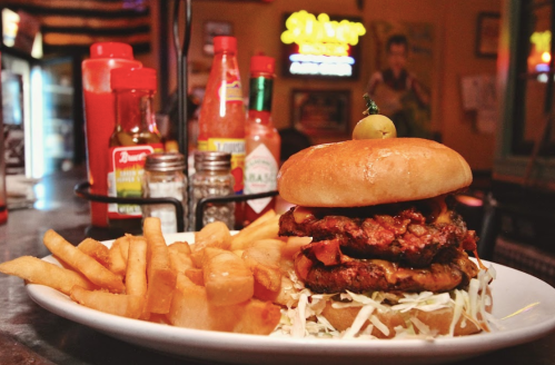 A stacked burger with fried chicken, cheese, and lettuce, served with crispy fries and condiments in the background.