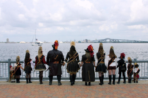 A group of people in pirate costumes stands by the water, facing a city skyline with a sailboat in the distance.