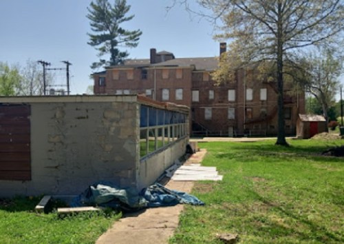 Abandoned building with a brick facade, surrounded by grass and trees, featuring a nearby structure with glass windows.