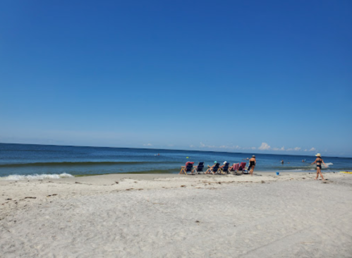 A sunny beach scene with people relaxing in chairs by the water, clear blue sky, and gentle waves.