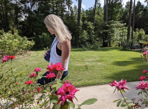 A woman with long blonde hair walks through a garden filled with pink roses, surrounded by greenery and trees.