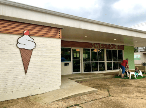 A quaint ice cream shop with a large cone sign, featuring a person and a dog outside on a grassy area.