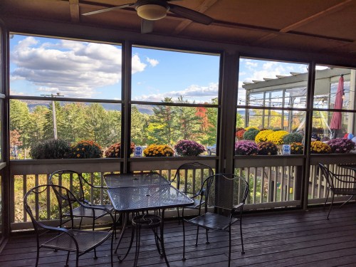 A screened porch with a table and chairs, overlooking colorful flower pots and a scenic view of trees and clouds.