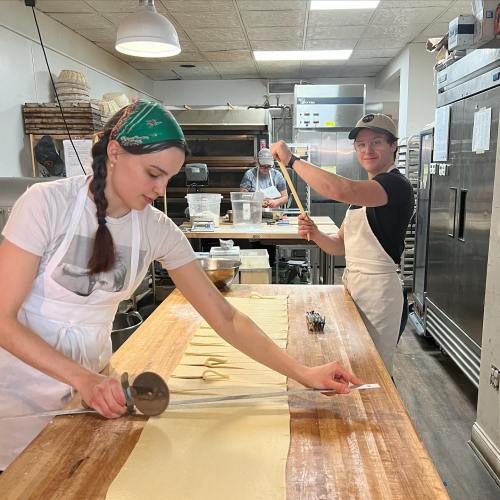 Two women in aprons prepare dough in a kitchen, one cutting pasta while the other stretches it on a wooden table.