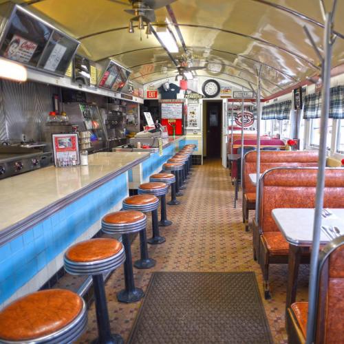 A retro diner interior featuring a long counter, bar stools, and booths, with vintage decor and a checkered floor.