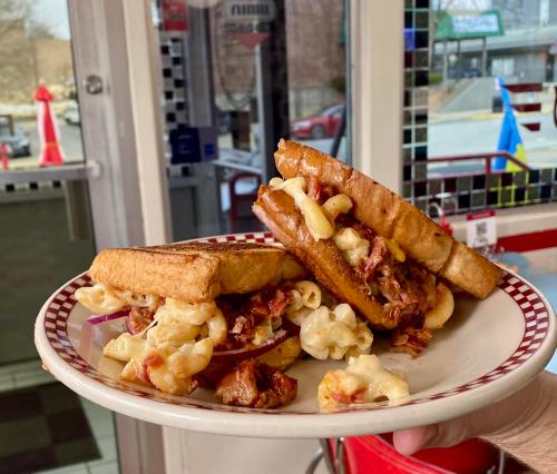 A plate holds two grilled cheese sandwiches filled with meat and topped with macaroni and cheese, set in a diner.