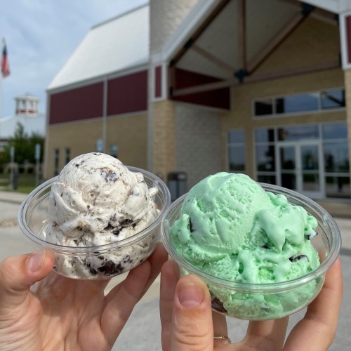 Two cups of ice cream held in hands: one with cookies and cream, the other mint green, in front of a building.
