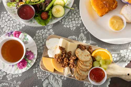 A wooden platter with assorted cheeses, nuts, crackers, and fruit, alongside a cup of tea and a salad on a lace tablecloth.