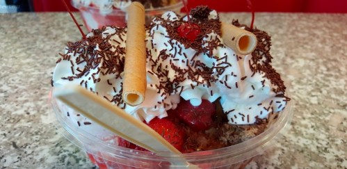 A close-up of a dessert cup topped with whipped cream, chocolate sprinkles, cherries, and rolled wafer cookies.