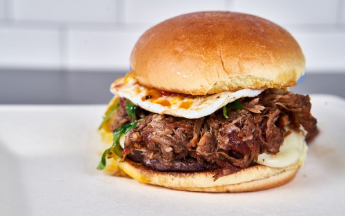 A hearty sandwich with pulled meat, melted cheese, a fried egg, and greens, served on a soft bun.