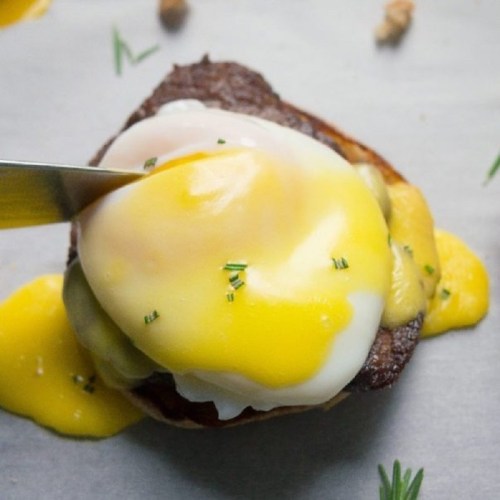 A close-up of a poached egg on a savory dish, with a knife cutting through the yolk, surrounded by herbs.