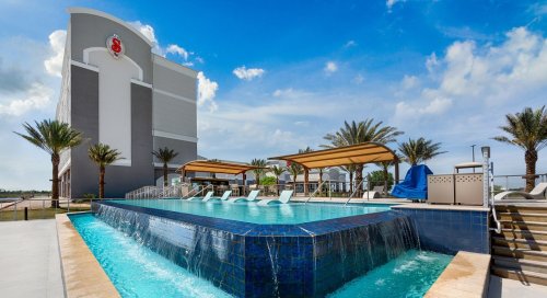 A modern hotel pool with lounge chairs, palm trees, and a clear blue sky in the background.