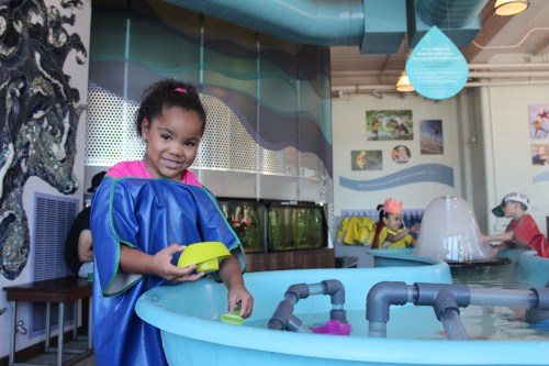 A smiling girl in a blue smock plays with colorful toys in a water activity area at a children's museum.