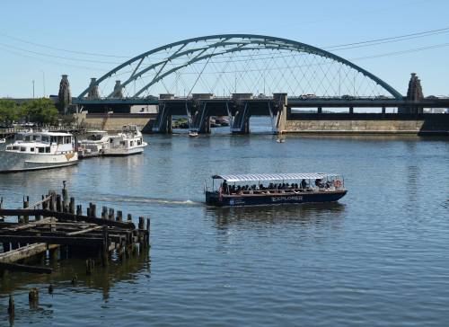 A boat named "Explorer" navigates a river under a large arch bridge, with moored boats in the foreground.