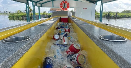 A long bar filled with ice and various cans of beverages, set in a waterfront location with greenery in the background.