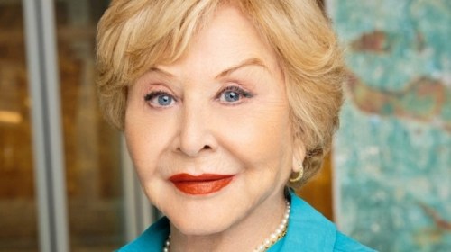 A smiling older woman with blonde hair, wearing a turquoise blouse and pearl necklace, against a textured background.