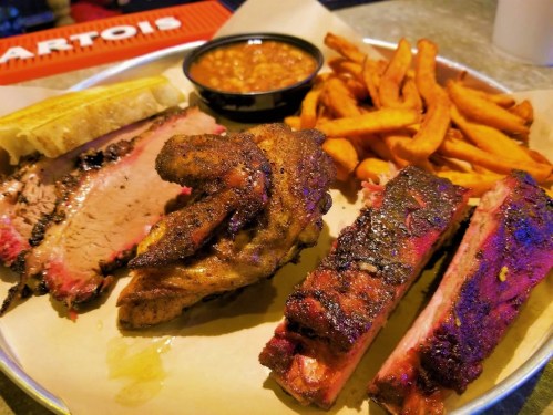 A platter of smoked meats including brisket, chicken wings, and ribs, served with sweet potato fries and baked beans.