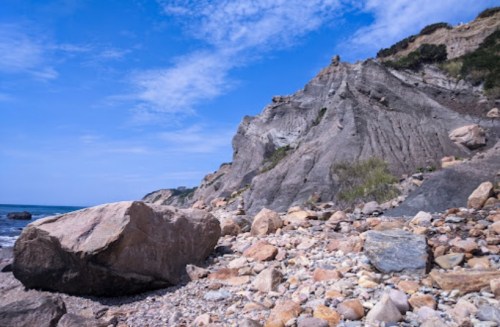 Rocky beach with large boulders and steep cliffs under a blue sky with scattered clouds.