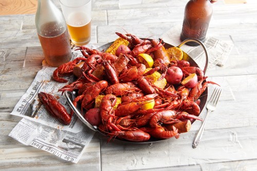 A large platter of boiled crawfish, potatoes, and lemon, with drinks and newspaper on a wooden table.