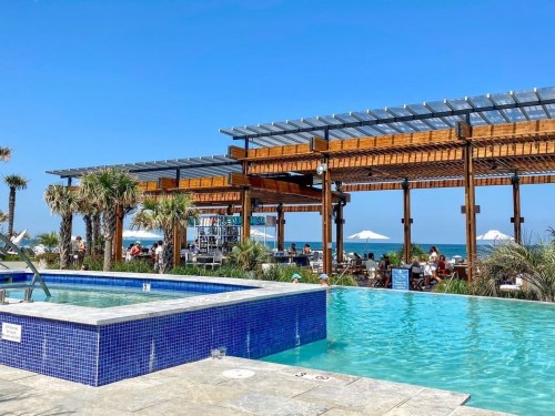 A sunny poolside view with palm trees, a wooden structure, and people dining near the beach.