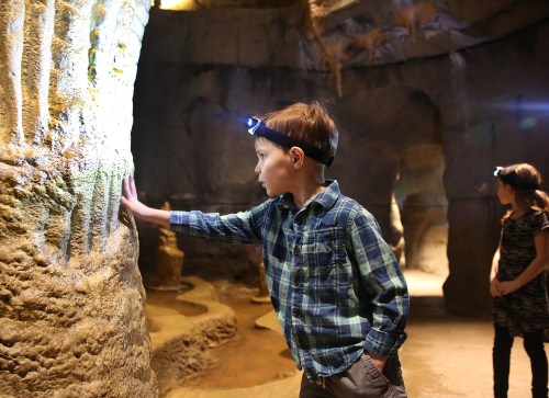 Two children explore a cave, one touching a rock formation while wearing a headlamp.