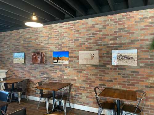 A brick wall in a cafe displays five nature-themed artworks, with wooden tables and metal chairs in the foreground.