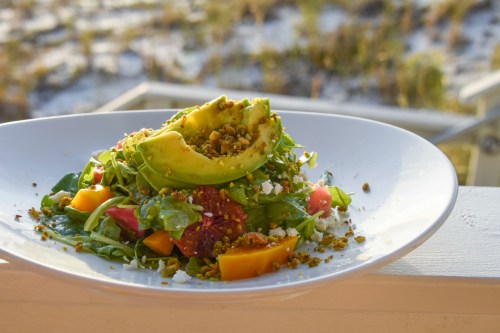 A vibrant salad with greens, avocado, tomatoes, and nuts, served on a white plate against a blurred outdoor background.
