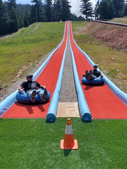 Two people sit on inflatable tubes at the top of a colorful slide, surrounded by trees and grass.