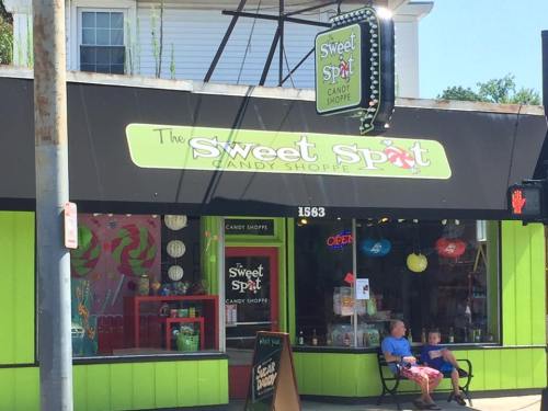 A colorful candy shop with a bright sign, featuring two children sitting outside and a variety of candy displayed.