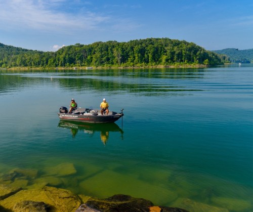 Two fishermen in a small boat on a calm lake, surrounded by lush green hills under a clear blue sky.