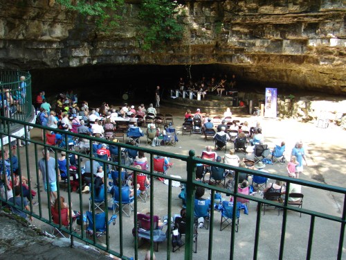 A crowd sits in chairs watching a performance in a cave-like setting, surrounded by greenery and rock formations.