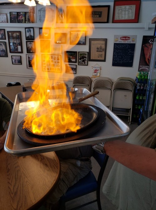 A person holds a tray with a flaming skillet, surrounded by a casual dining setting. Flames rise dramatically.