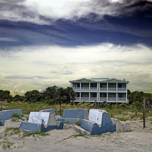 A beach scene featuring a blue and white building in the background, with concrete structures in the foreground and cloudy skies.