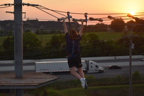 A person hangs from a cable at sunset, with a highway and trees in the background.