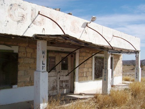 Abandoned building with a weathered facade, overgrown grass, and faded signs for cigarettes and gifts.