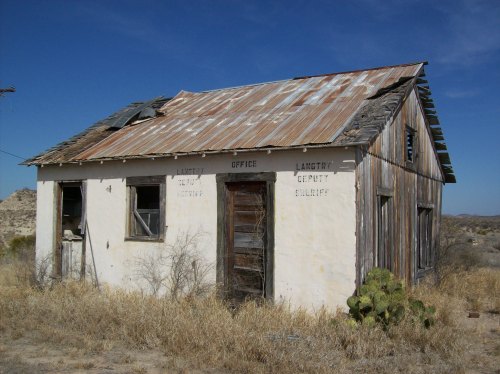 Abandoned building with a rusty roof, labeled "Office" and "Deputy Sheriff," surrounded by dry grass and desert landscape.