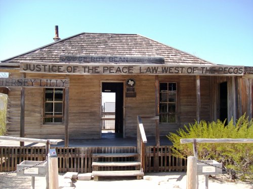 Historic wooden building with a sign reading "Justice of the Peace Law West of the Pecos," surrounded by desert vegetation.