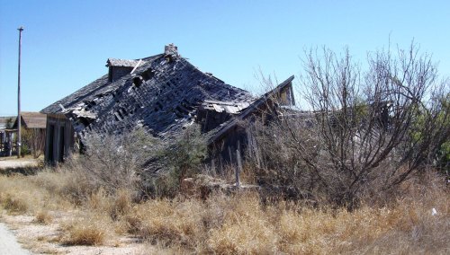 An abandoned, dilapidated building with a sagging roof, surrounded by dry grass and sparse vegetation under a clear blue sky.