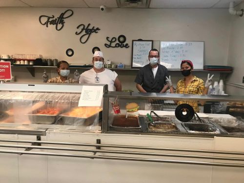 Four people in masks stand behind a food counter with trays of food, smiling in a restaurant setting.