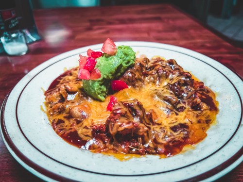 A plate of enchiladas topped with melted cheese, guacamole, and diced tomatoes, served on a wooden table.